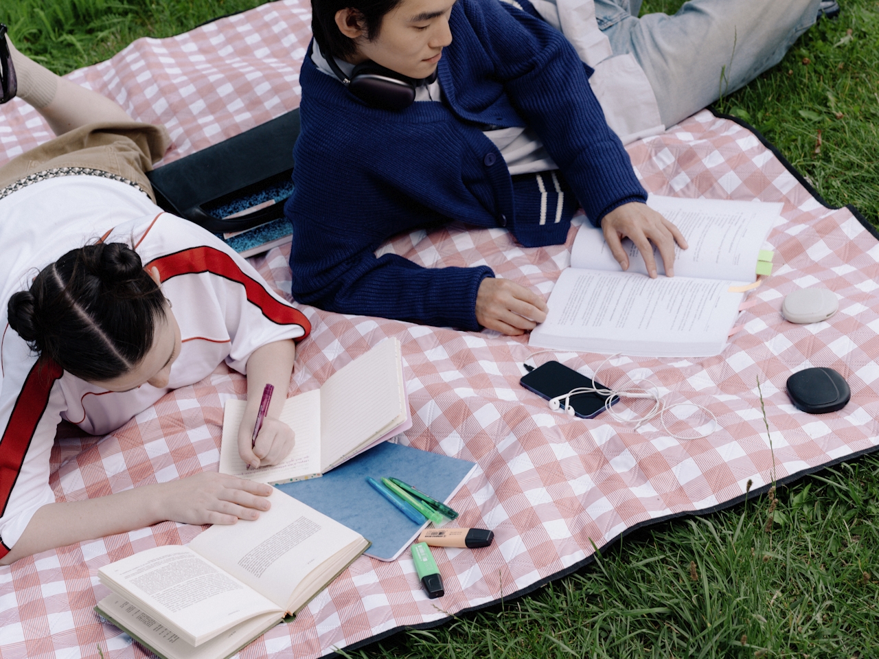 Two students studying outdoors on a pink and white checkered blanket with open books, notebooks, and pens.