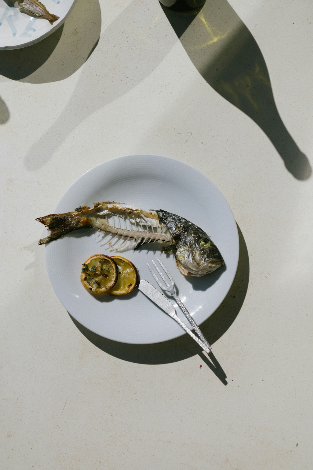 A white plate with a partially eaten fish skeleton, two slices of grilled lemon, and a fork and knife on a light-colored table with bottle shadows.