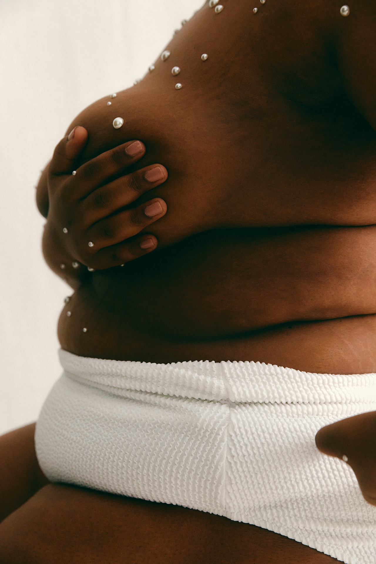 Close-up of a person with dark skin wearing white textured bottoms and holding their breast adorned with small adhesive pearls.