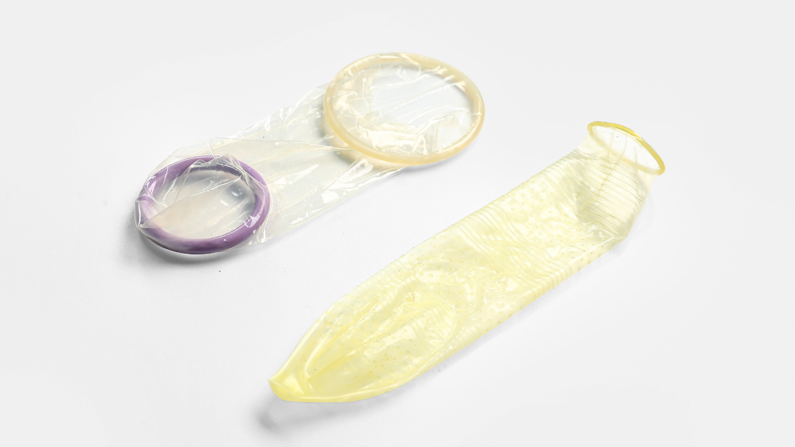 Two unrolled condoms on a white surface, one with a purple ring and the other yellow with textured dots and lines.