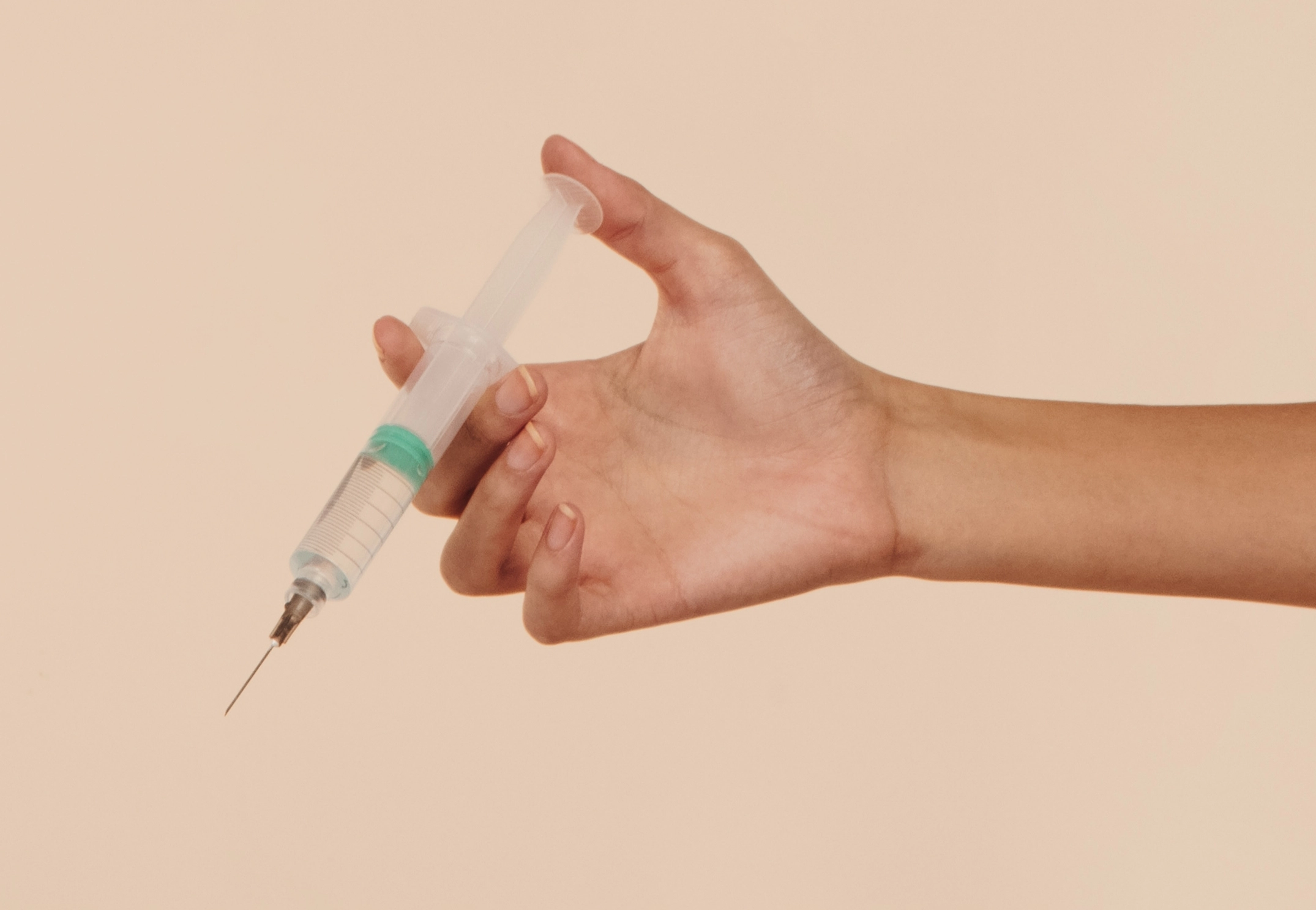 Hand holding a syringe with a clear barrel and a green stopper against a beige background.