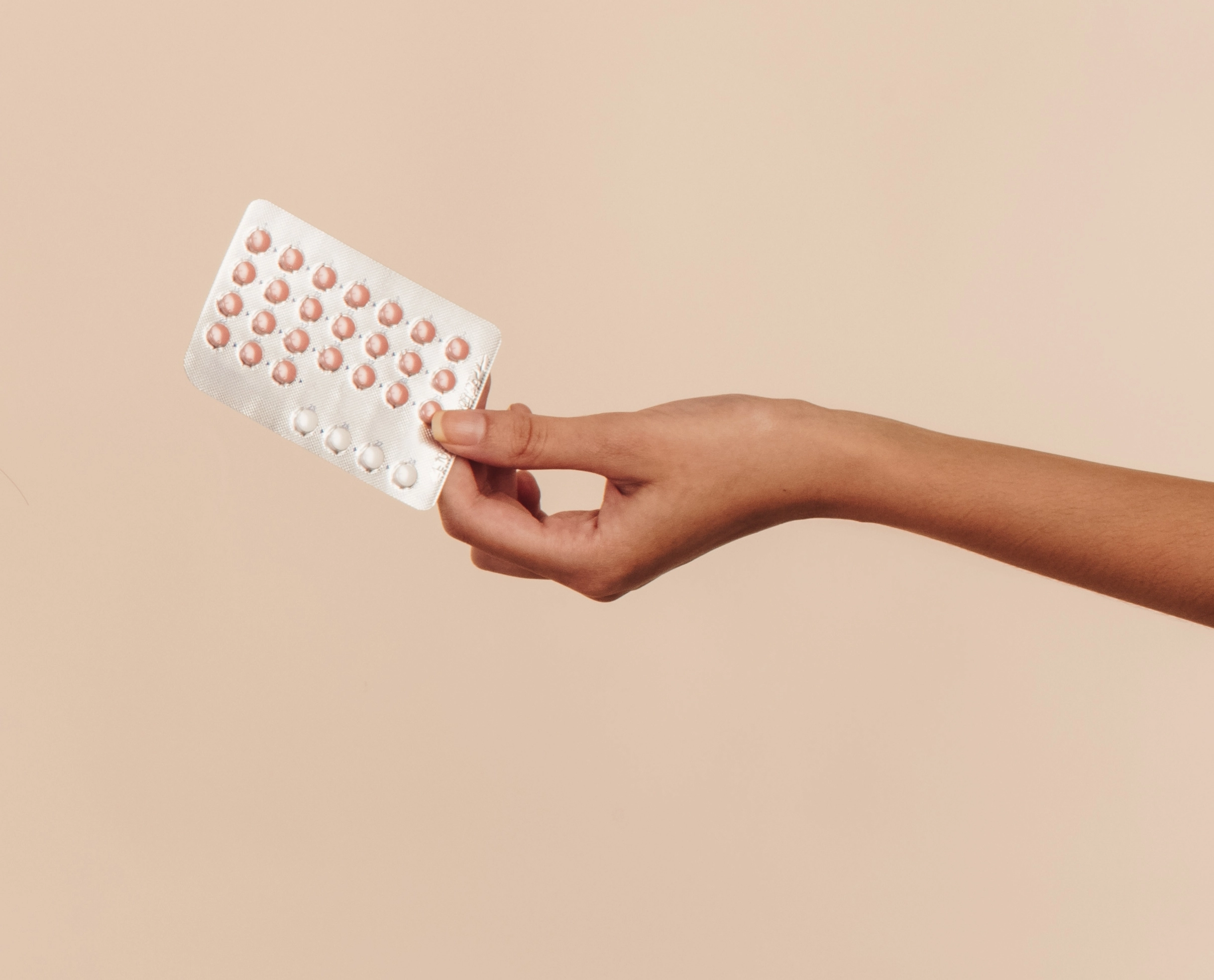 Hand holding a blister pack of birth control pills with pink and white tablets against a beige background.