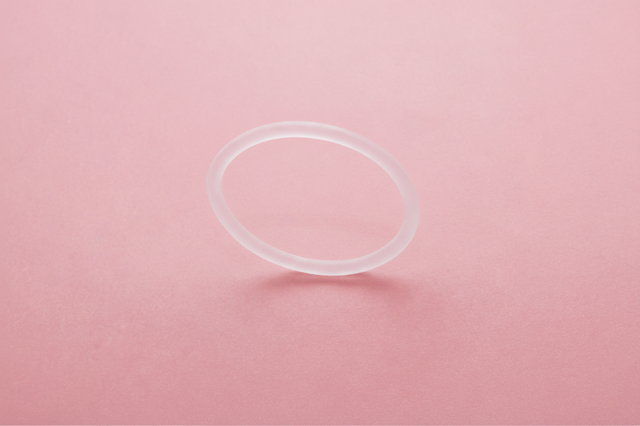 Translucent circular vaginal ring on a pink background, casting a soft shadow.