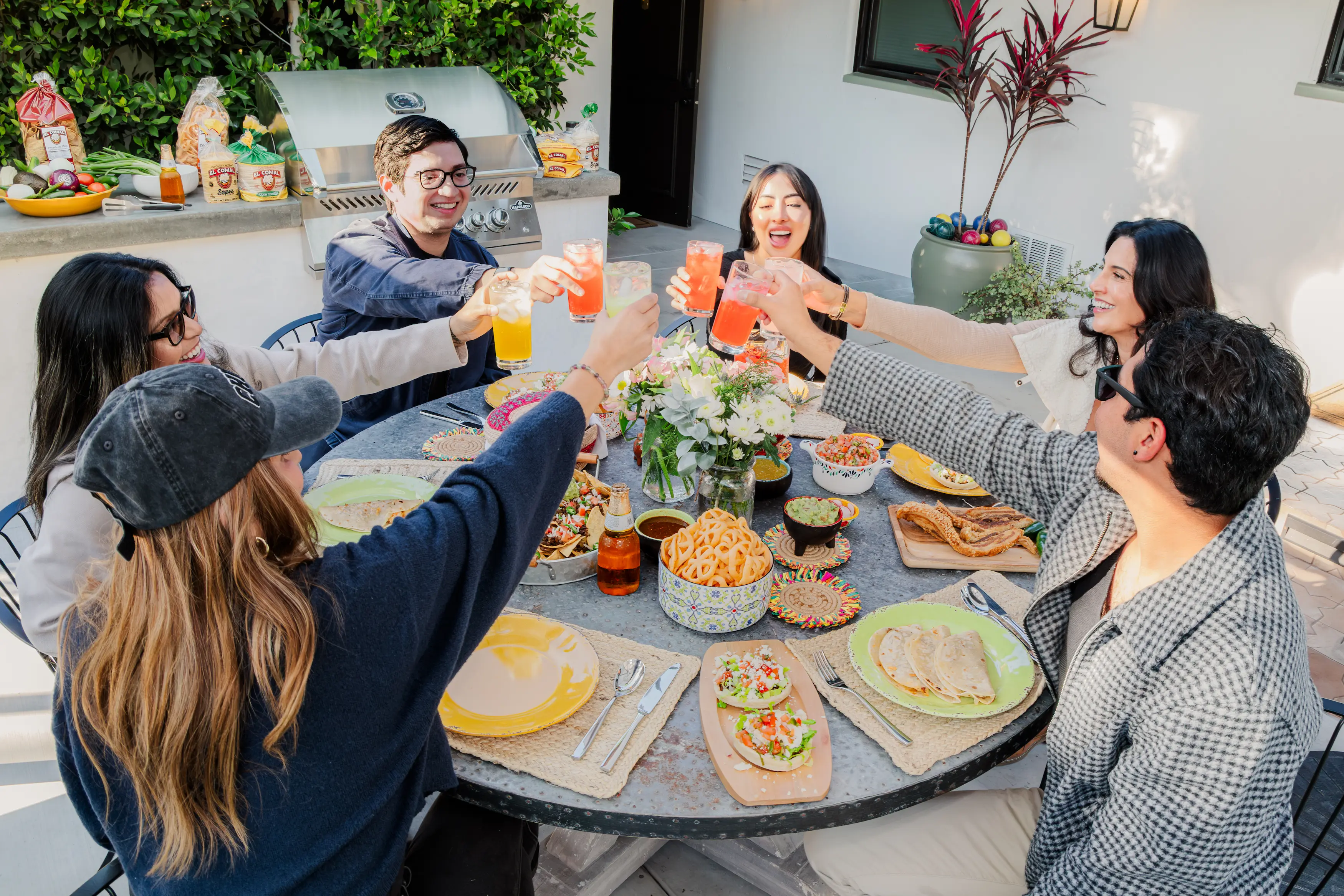 Friends raising glasses in a toast around a patio table filled with tacos, quesadillas, guacamole, chips, and Mexican dishes, with El Comal products and a barbecue grill in the background