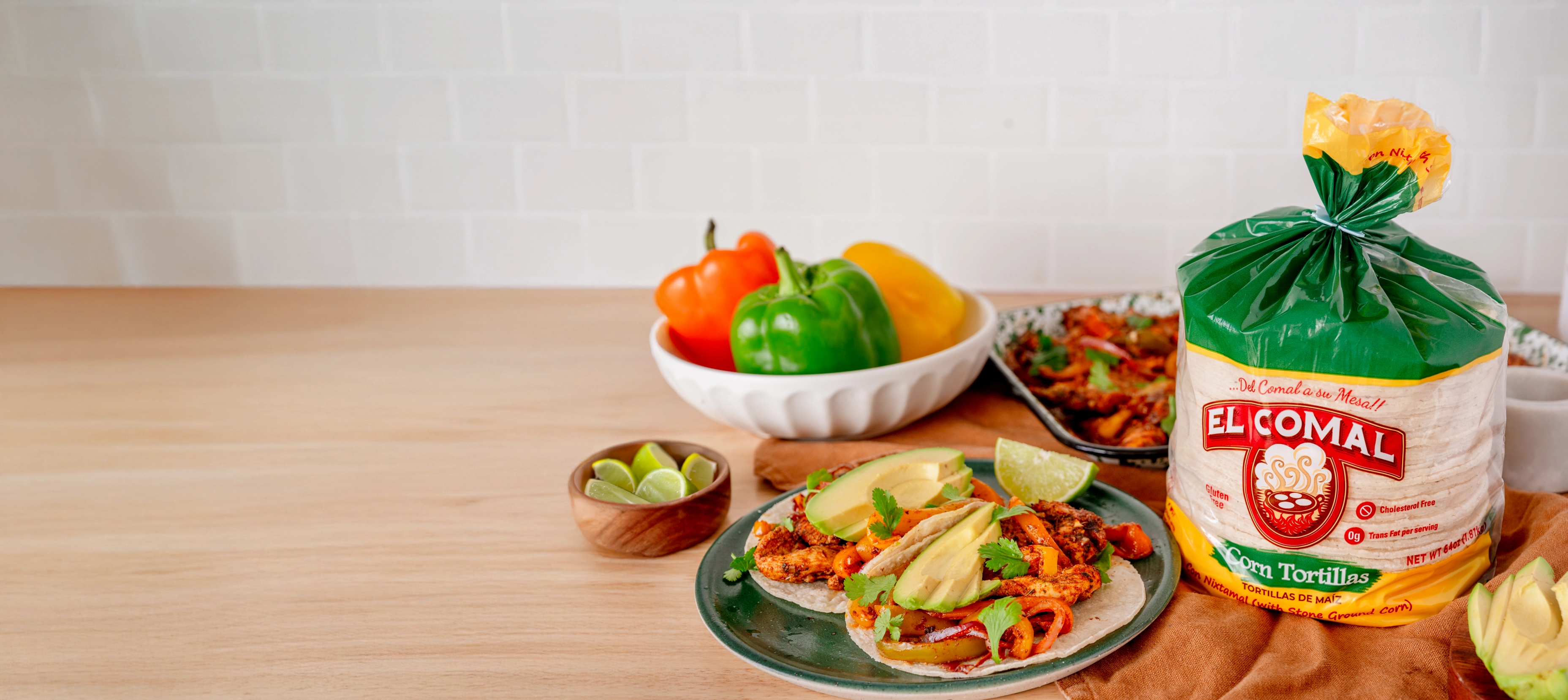 Chicken fajita tacos with avocado slices on a plate, surrounded by fresh bell peppers in a bowl, lime wedges, and a bag of El Comal corn tortillas on a wooden cutting board