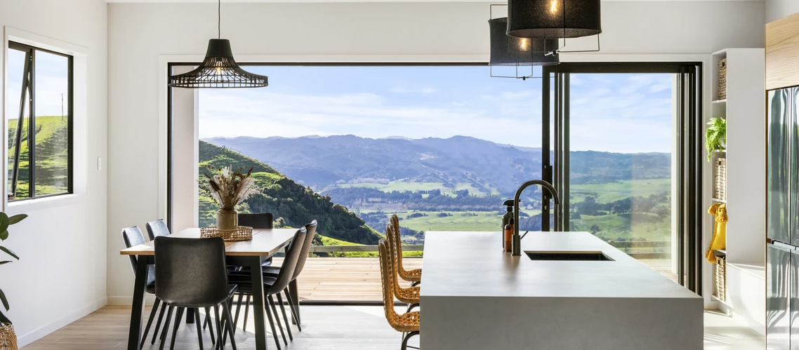Modern open-plan kitchen and dining area with floor-to-ceiling aluminium sliding doors by Rodney Aluminium, showcasing seamless indoor-outdoor living and a rural Mangawhai landscape.