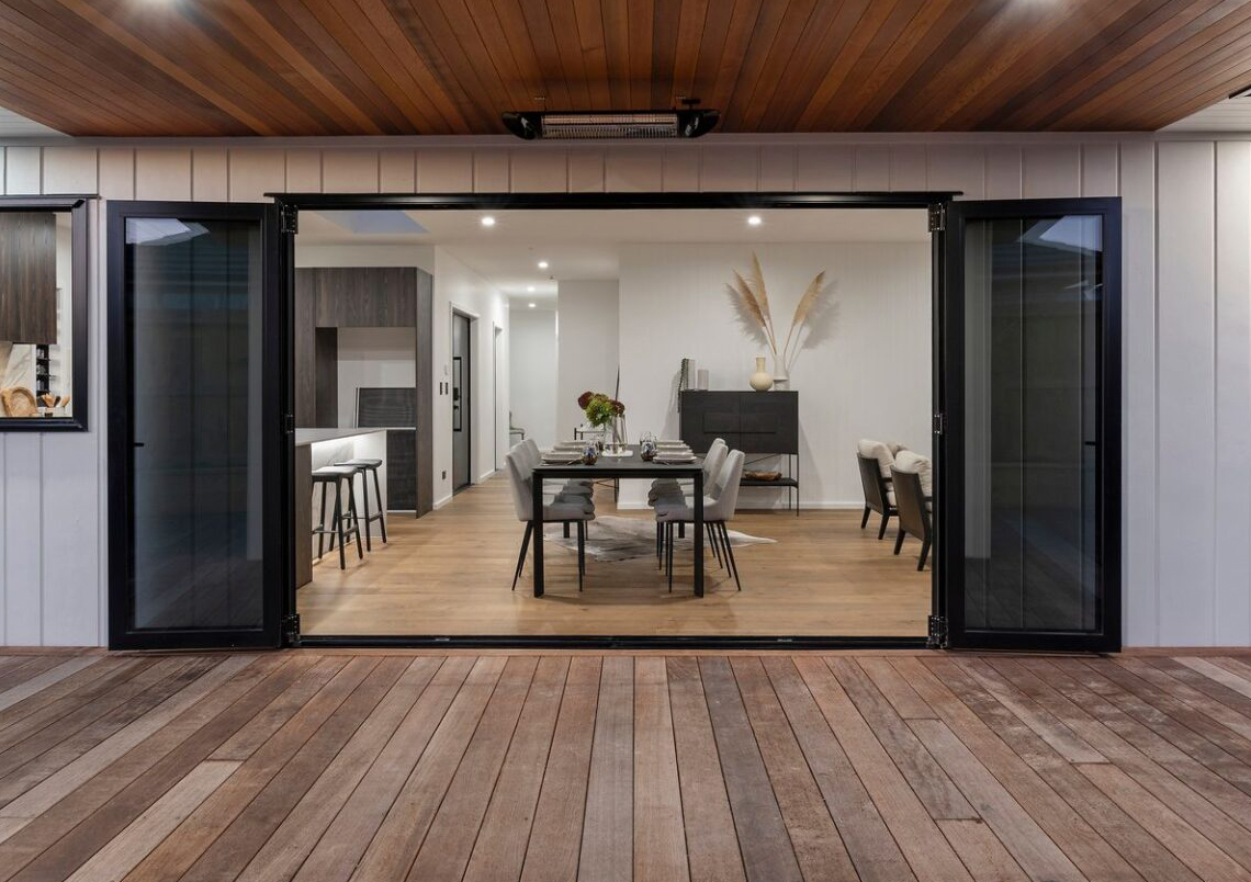 Modern indoor-outdoor dining area with black aluminium bifold doors by Rodney Aluminium, fully opened to connect the interior living space with the timber deck in a contemporary Mangawhai home.