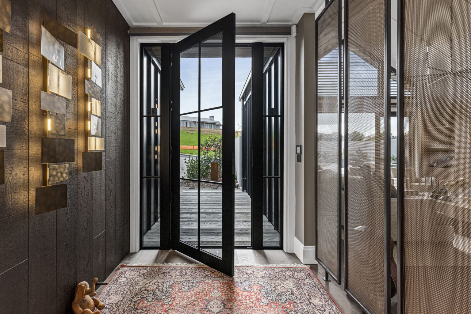 Contemporary entryway featuring a black aluminium pivot door by Rodney Aluminium, with sidelights and modern detailing, opening to an exterior timber deck.