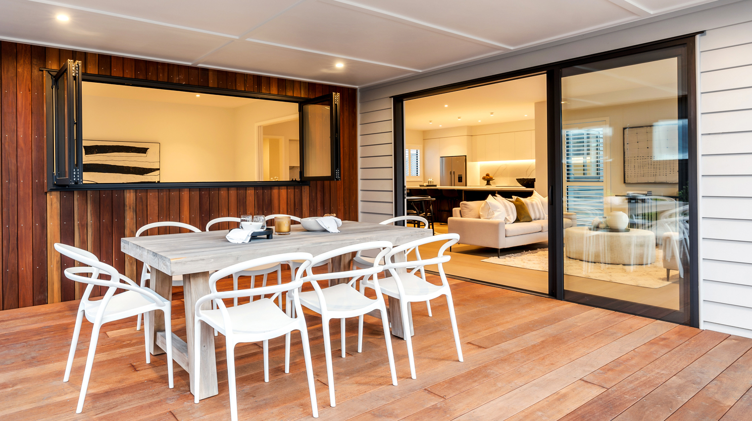 Outdoor dining area with custom black aluminium sliding door and bifold servery window by Rodney Aluminium, connecting indoor kitchen and living space to the timber deck in a modern Warkworth home.