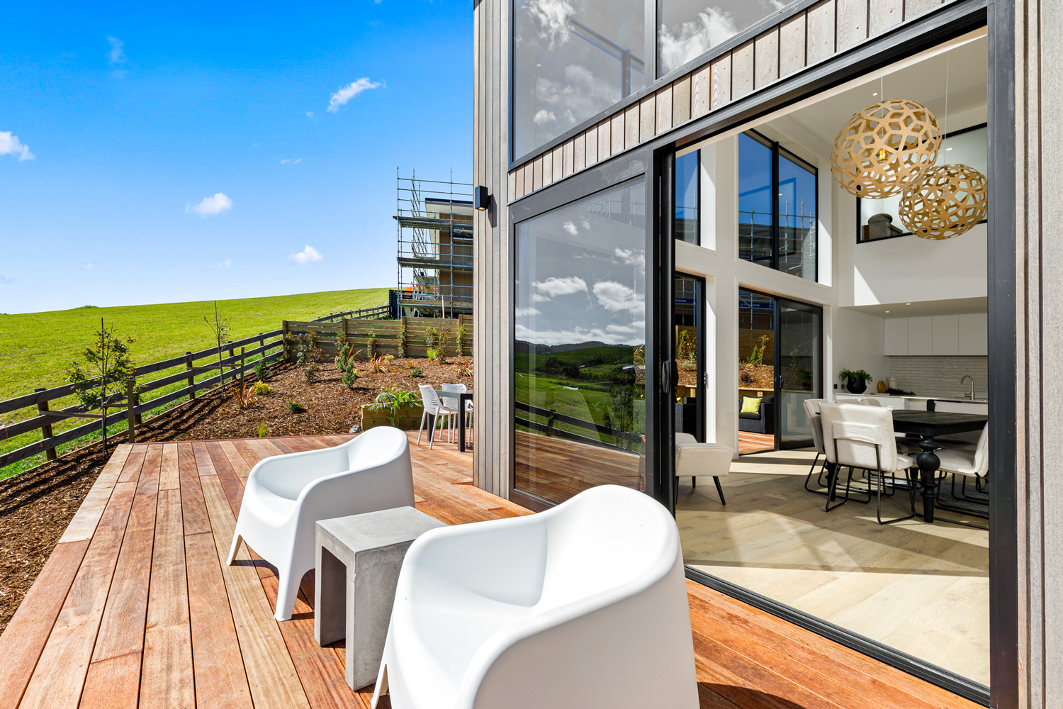 Spacious timber deck with black aluminium sliding and stacking glass doors by Rodney Aluminium, seamlessly connecting the open-plan kitchen and dining area to the outdoors in a modern Matakana home.