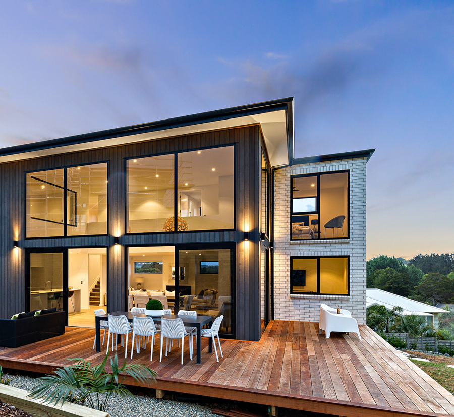 Modern two-story home at dusk featuring large aluminium-framed windows, timber decking, and outdoor dining area, showcasing contemporary architectural design in New Zealand.