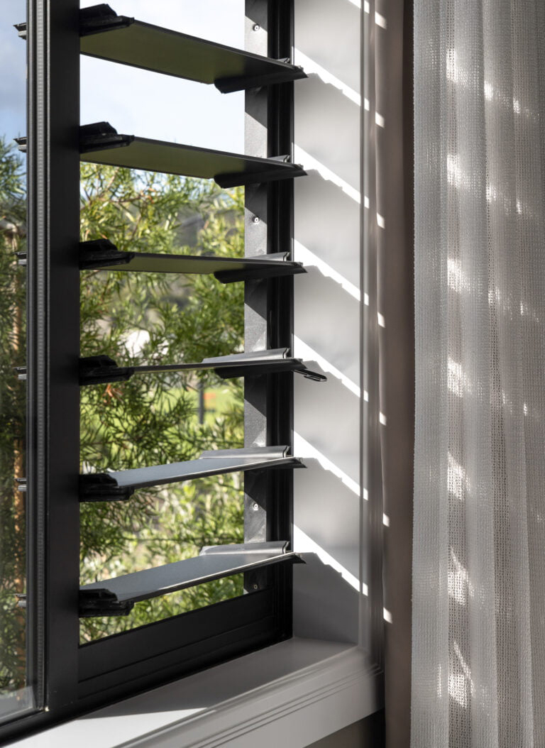 Close-up of modern aluminium louvre window partially open, with natural light casting shadows on white walls and sheer curtains, showcasing ventilation and architectural detail.