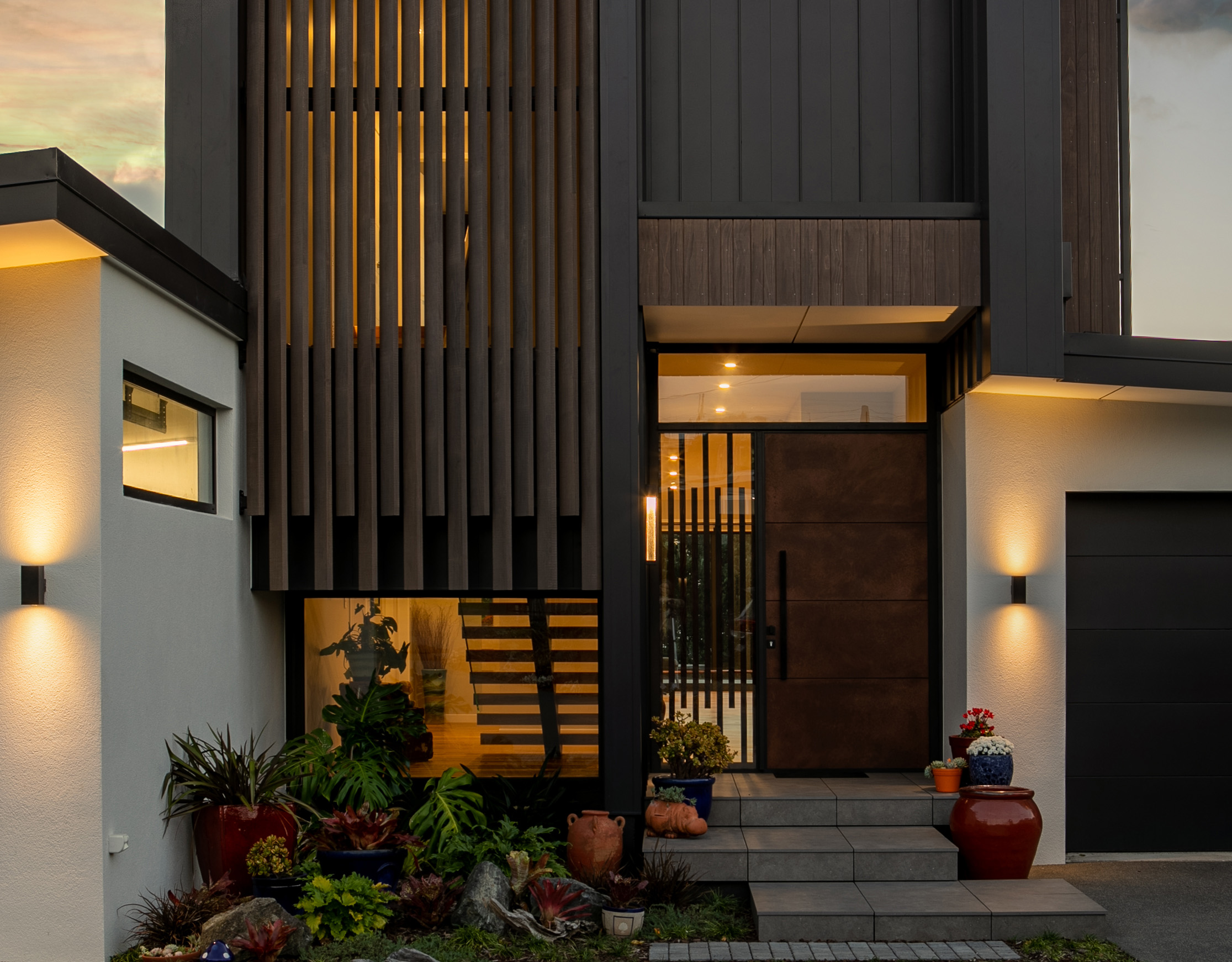 Contemporary front entrance featuring thermal aluminium joinery, large pivot door, vertical timber slats, and exterior wall lighting, with modern landscaping and potted plants, Warkworth, New Zealand.