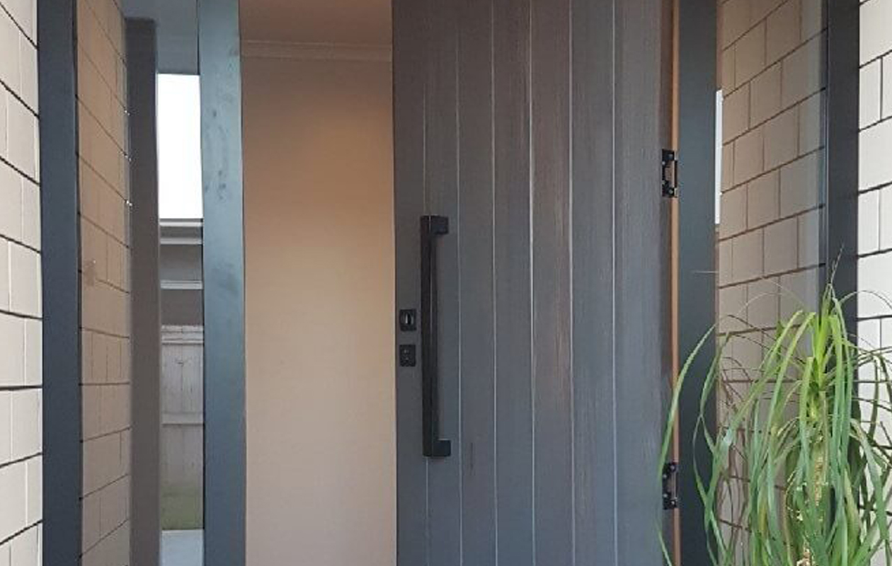 Modern front entry door with vertical panel design and matte black pull handle, installed by Rodney Aluminium, framed by brick cladding and side glazing.