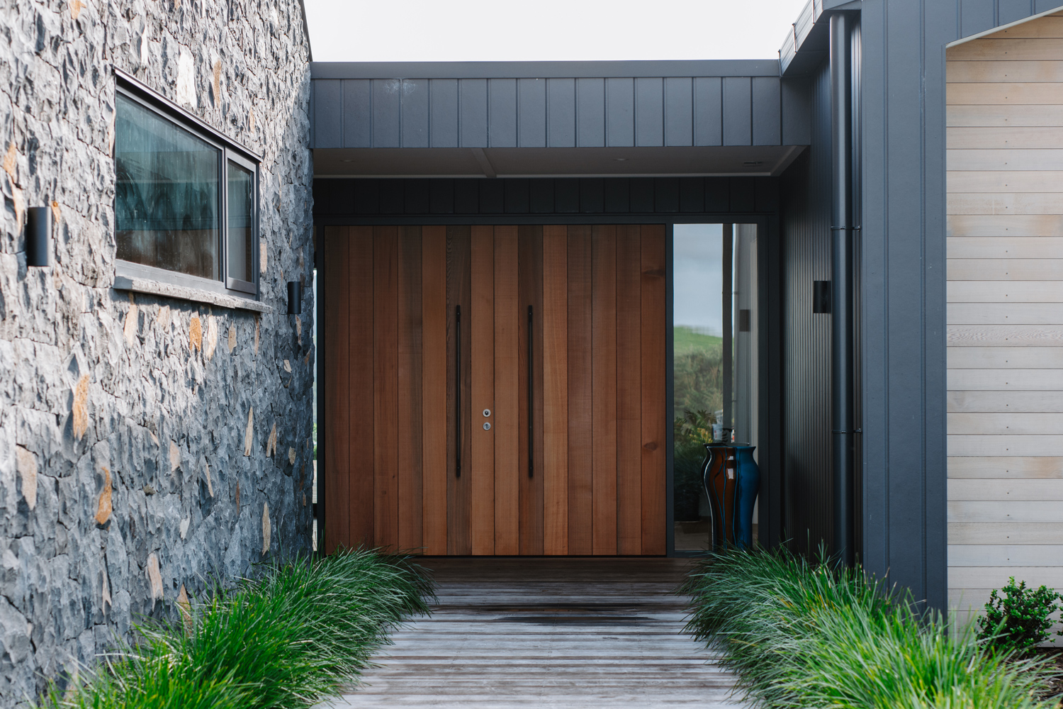 Modern front entrance with vertical timber double doors, surrounded by dark metal cladding and stone walls, featuring a narrow window and landscaped walkway.