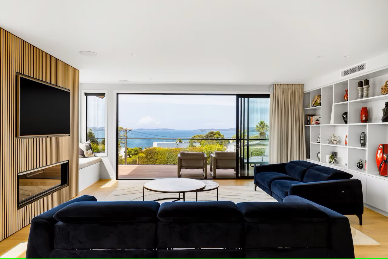 Aluminium sliding doors in a modern Matakana home, opening to a coastal view from a contemporary living room with timber paneling and custom shelving