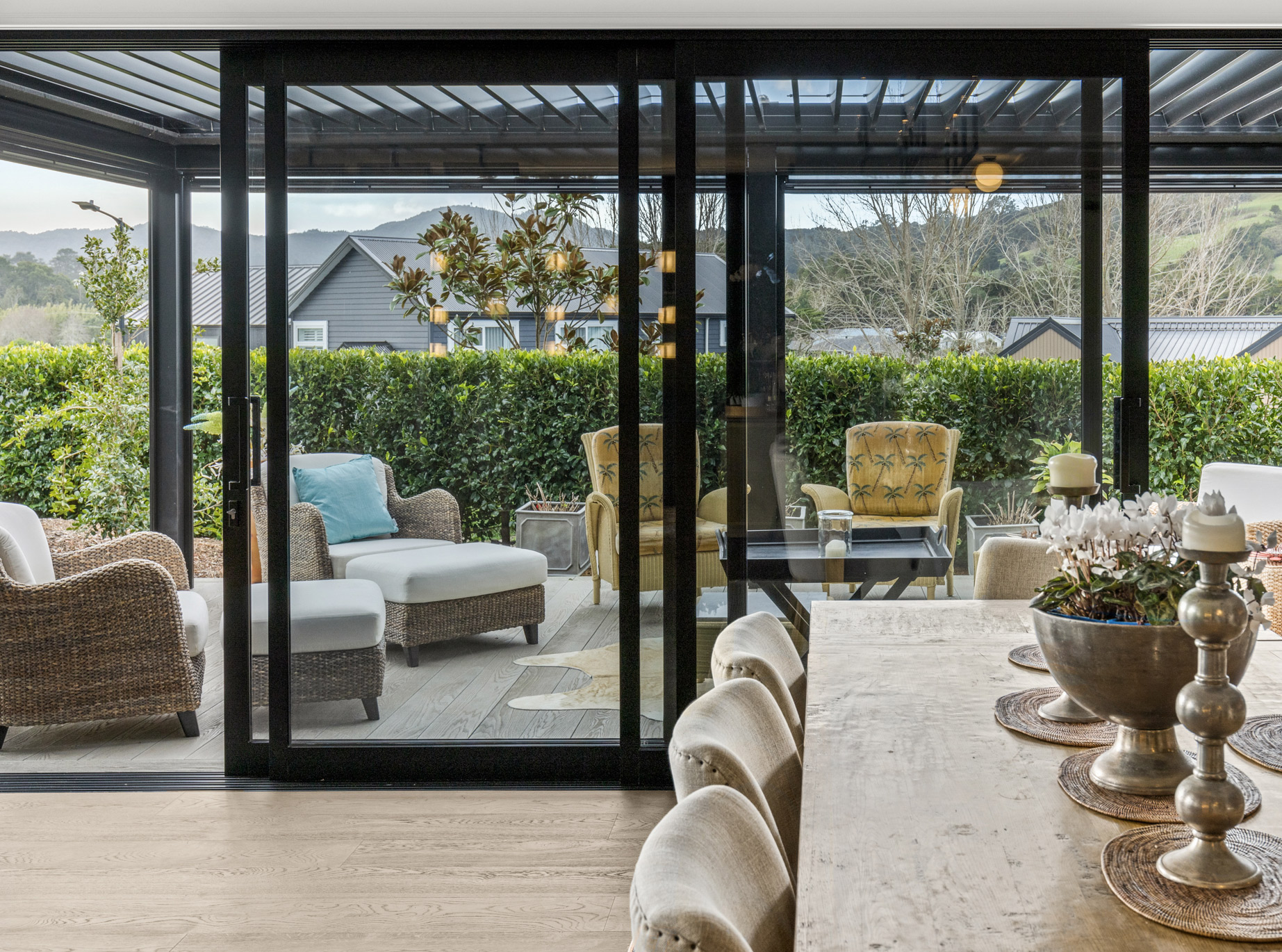 Modern aluminum sliding glass doors open onto a covered outdoor patio with wicker and upholstered seating, showcasing custom joinery and scenic rural views in Puhoi, New Zealand.