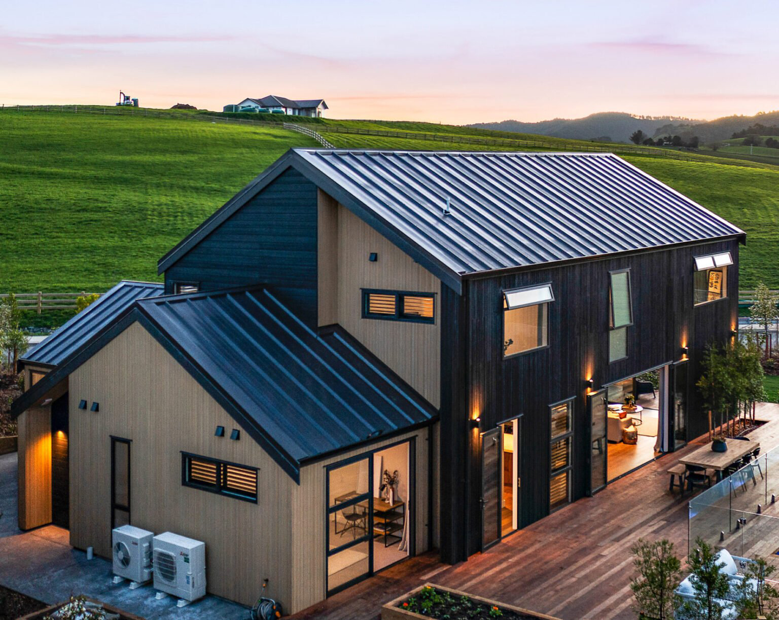 Contemporary rural home in Mangawhai featuring dark vertical cladding, aluminum windows and doors, and a spacious timber deck overlooking rolling green hills at sunset.