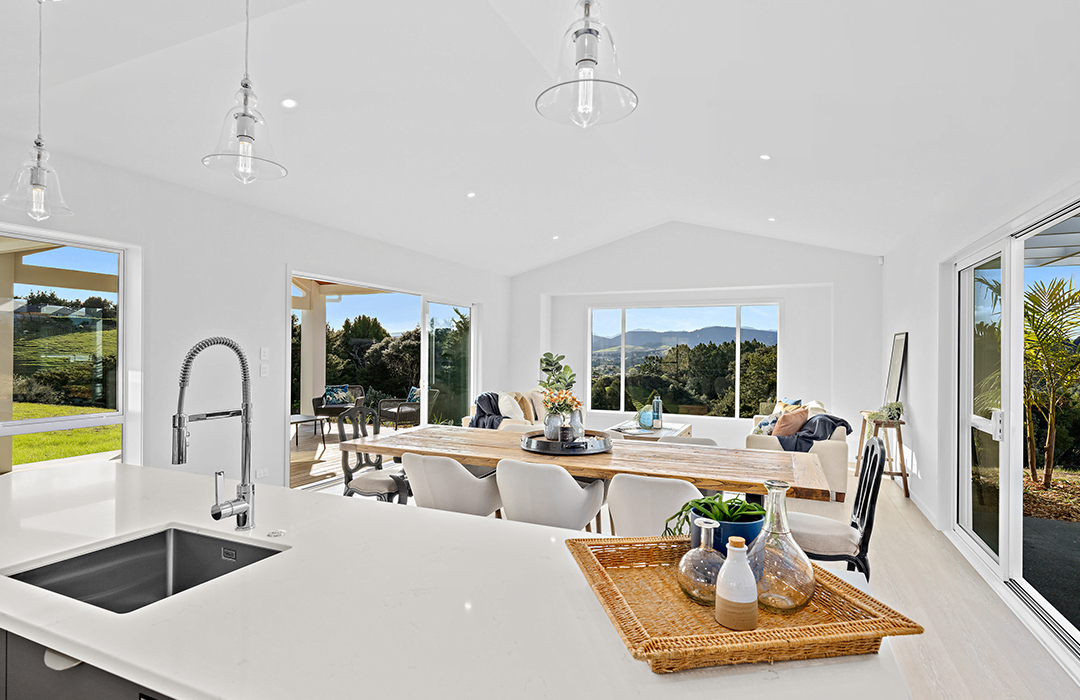 Bright, modern open-plan kitchen and dining area with large aluminum sliding and fixed windows, showcasing expansive rural views and custom joinery in a New Zealand home.