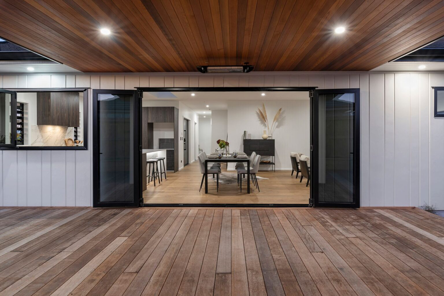 Evening view of a contemporary dining and kitchen area with fully opened aluminum bifold doors, showcasing seamless indoor-outdoor flow, timber decking, and custom joinery by Rodney Aluminium.