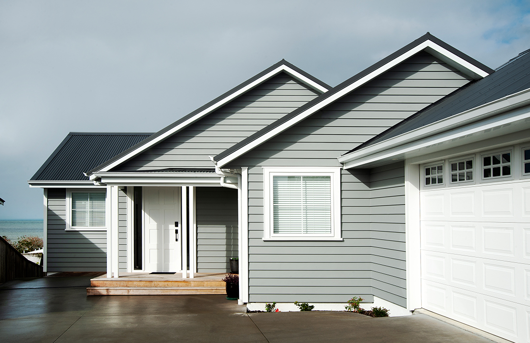 Front view of a modern coastal-style home with grey weatherboard cladding, white aluminium entry door, and matching garage door – showcasing sleek, durable exterior finishes by Rodney Aluminium