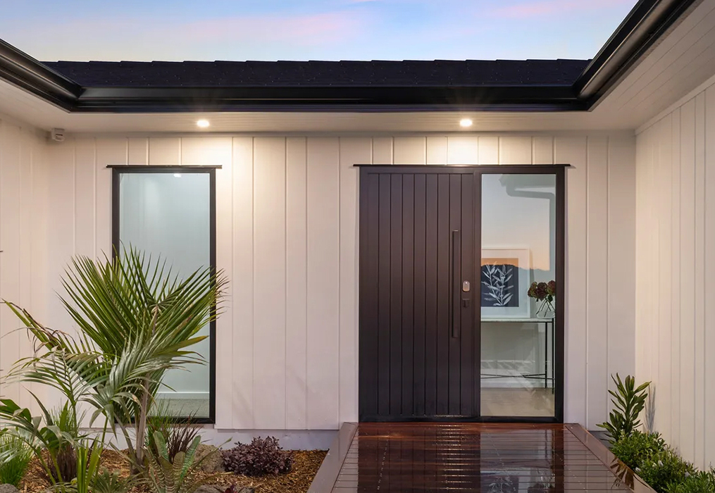 Contemporary home entrance featuring a sleek black aluminium tongue and groove front door with sidelights, set against vertical white cladding – supplied by Rodney Aluminium in Warkworth