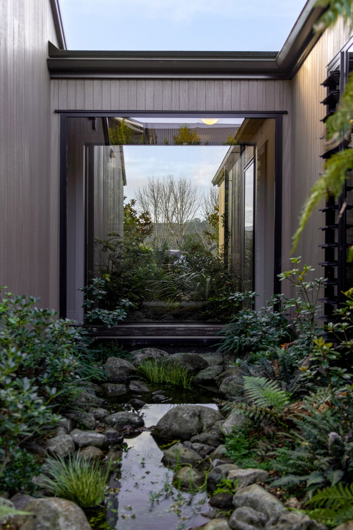 Frameless glass window feature looking out onto a landscaped rock stream and native garden between two modern cladded buildings – Rodney Aluminium NZ.