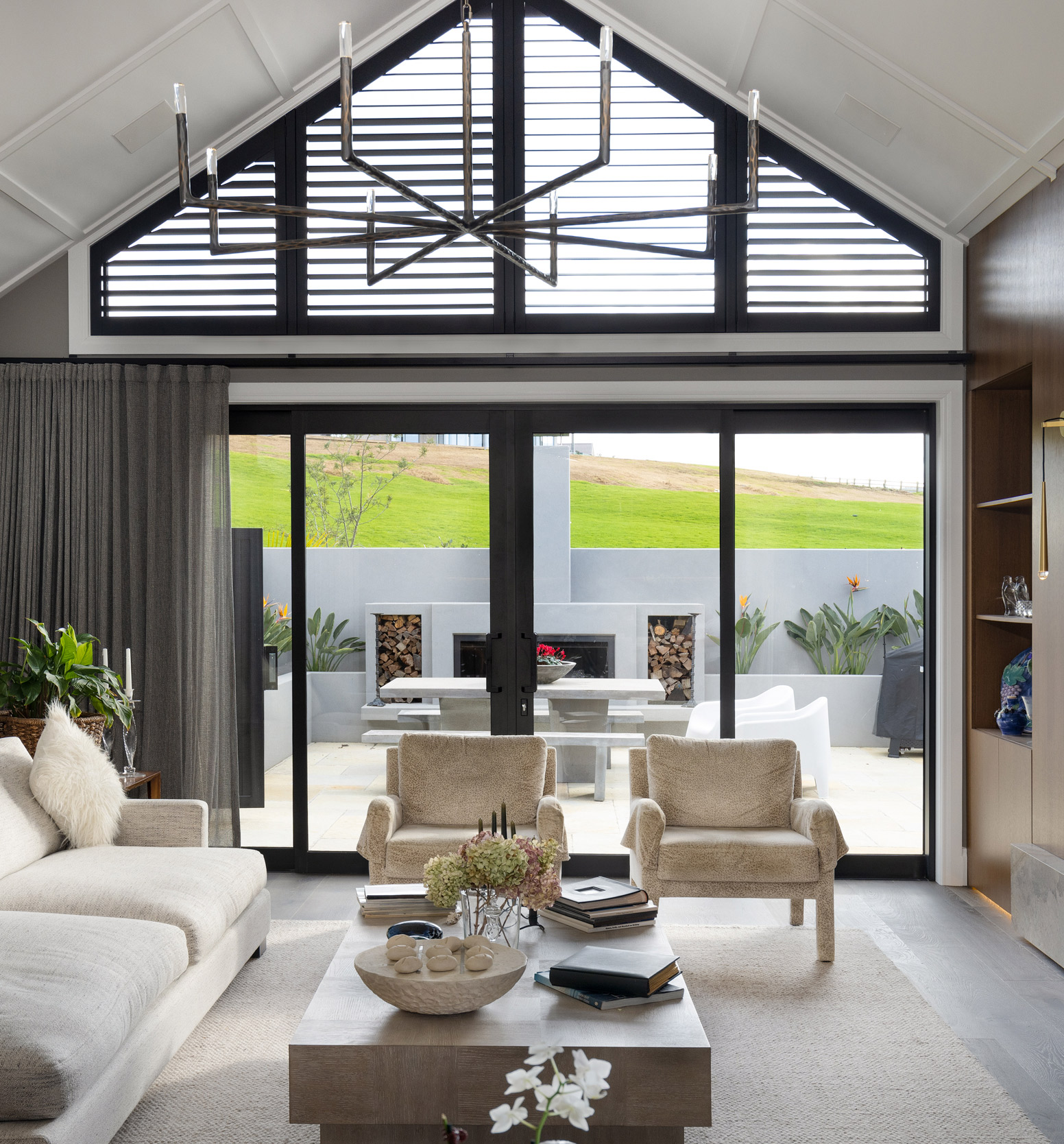 Modern living room with large black-framed sliding doors and gable windows showcasing premium aluminium joinery, opening to a landscaped outdoor area with built-in fireplace and seating.