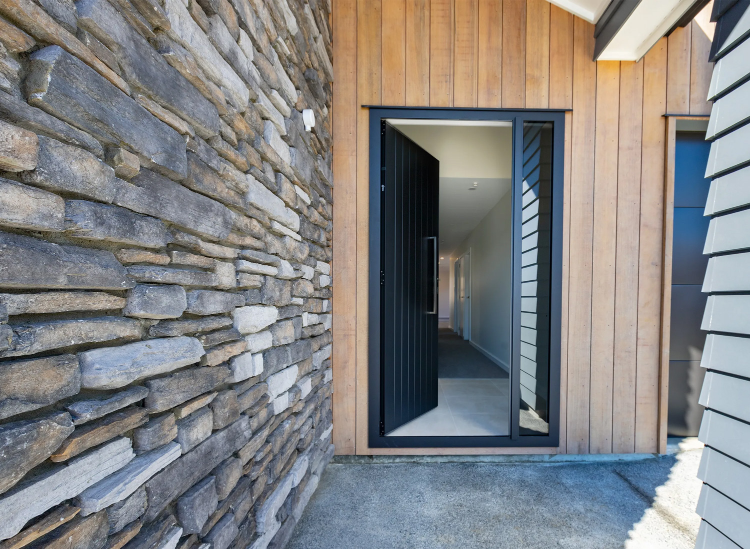 Modern black aluminium tongue-and-groove front door with sidelights, set in timber and schist exterior cladding, showcasing contemporary entry joinery design.