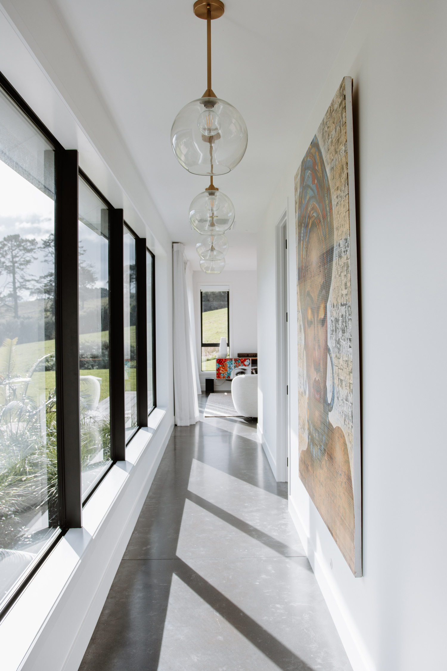 Bright hallway with black-framed aluminium windows showcasing expansive rural views, modern pendant lighting, and polished concrete floors in a contemporary Orewa home.