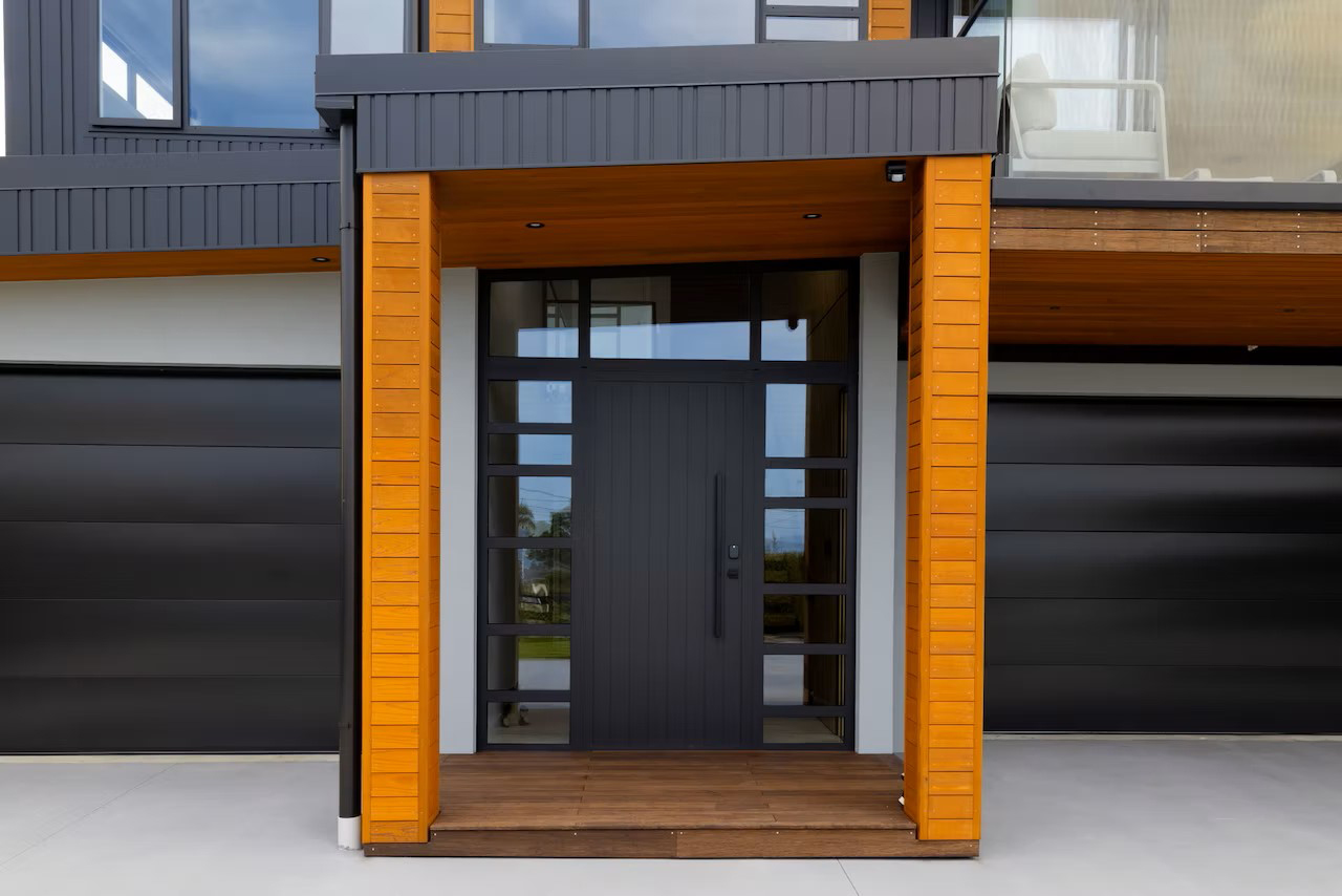 Contemporary front entrance with black aluminium tongue-and-groove door and sidelights, framed by vertical timber cladding and covered portico in a modern Silverdale home.