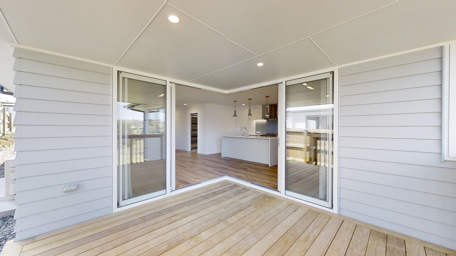 Open-plan home with corner-stacked, pillarless sliding glass doors in white aluminium frames, creating seamless indoor-outdoor flow onto a timber deck.