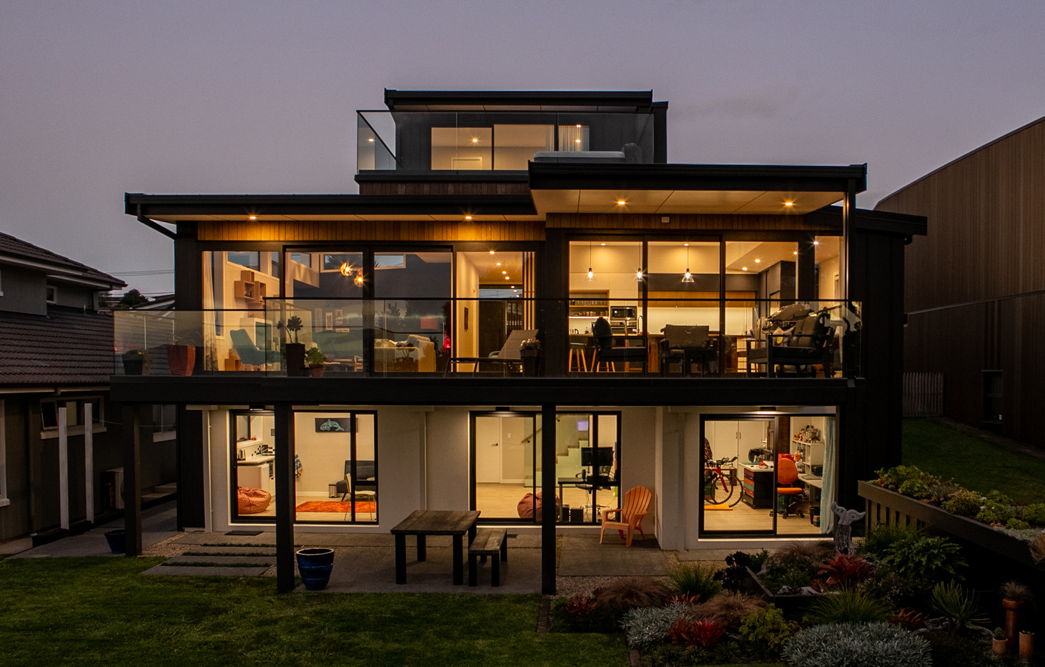 Three-level modern home at dusk featuring floor-to-ceiling aluminium joinery, glass balustrades, and expansive sliding doors, showcasing premium joinery by Rodney Aluminium in Tindalls Beach.