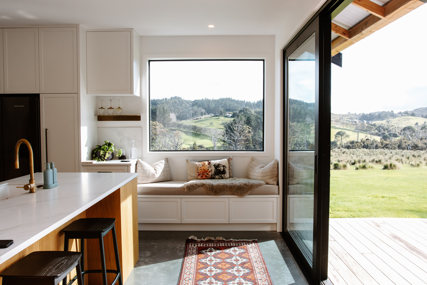 Bright kitchen and window seat with large fixed picture window and sliding door in black aluminium joinery, offering uninterrupted rural views in a Warkworth home.