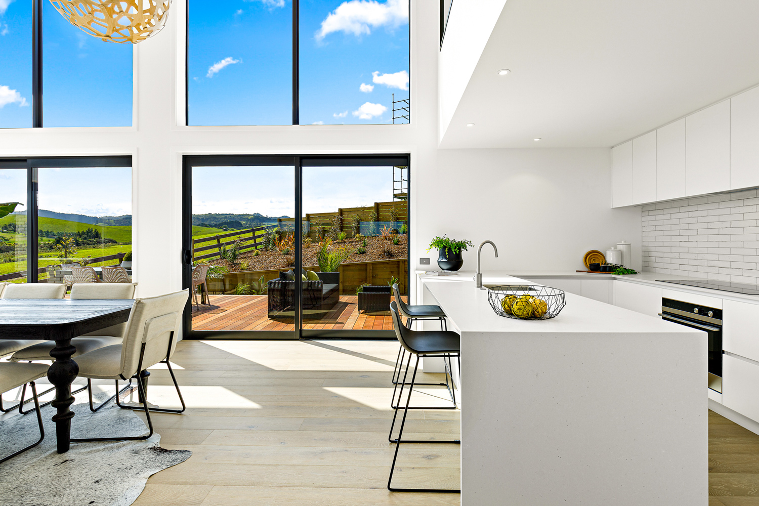 Bright contemporary kitchen and dining area with floor-to-ceiling aluminium windows and sliding doors, opening to a timber deck with scenic rural views in Kaiwaka.