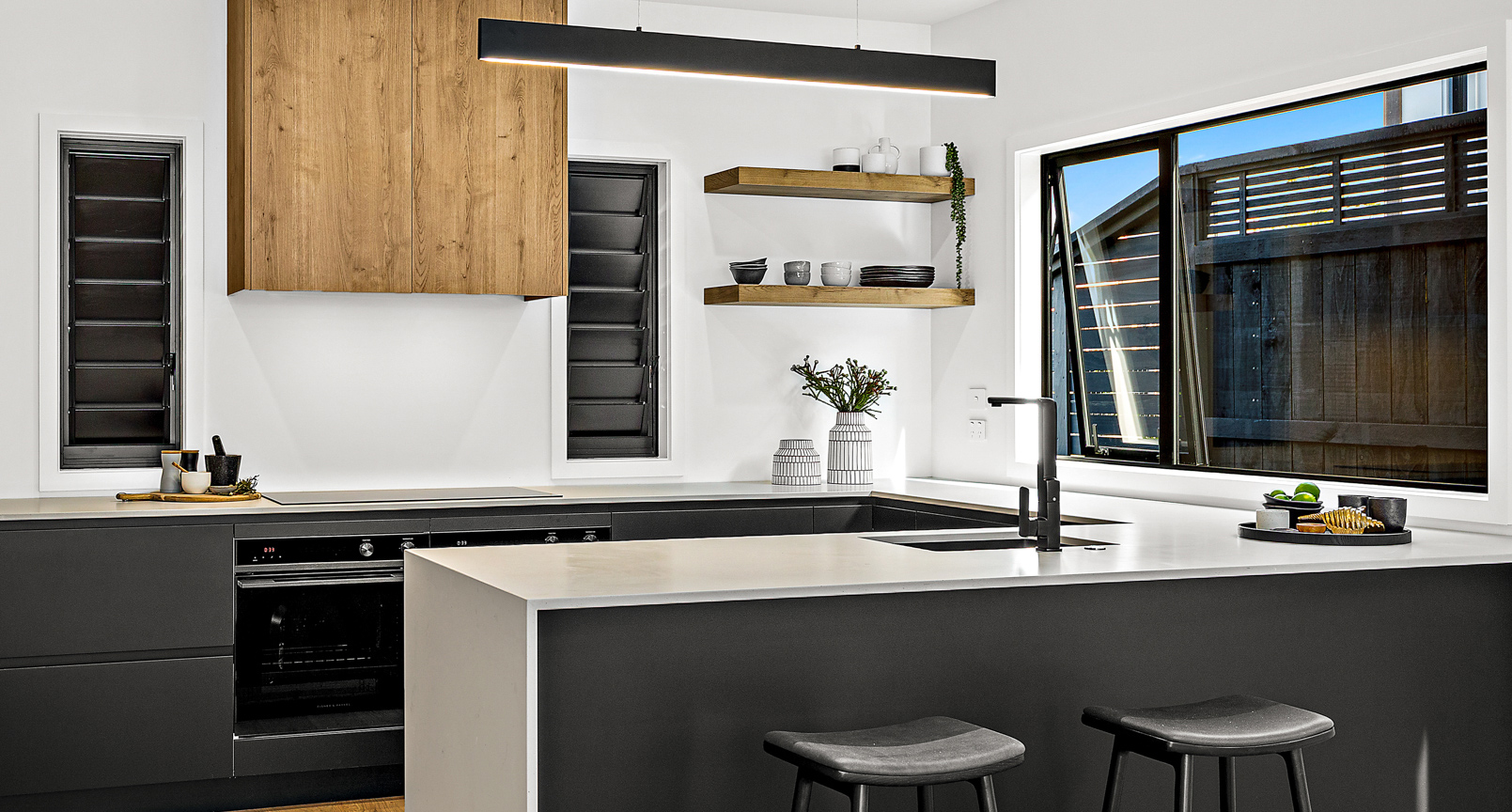 Sleek modern kitchen with black aluminium awning and louvre windows, matte cabinetry, wood accents, and white benchtops in a contemporary Leigh home.
