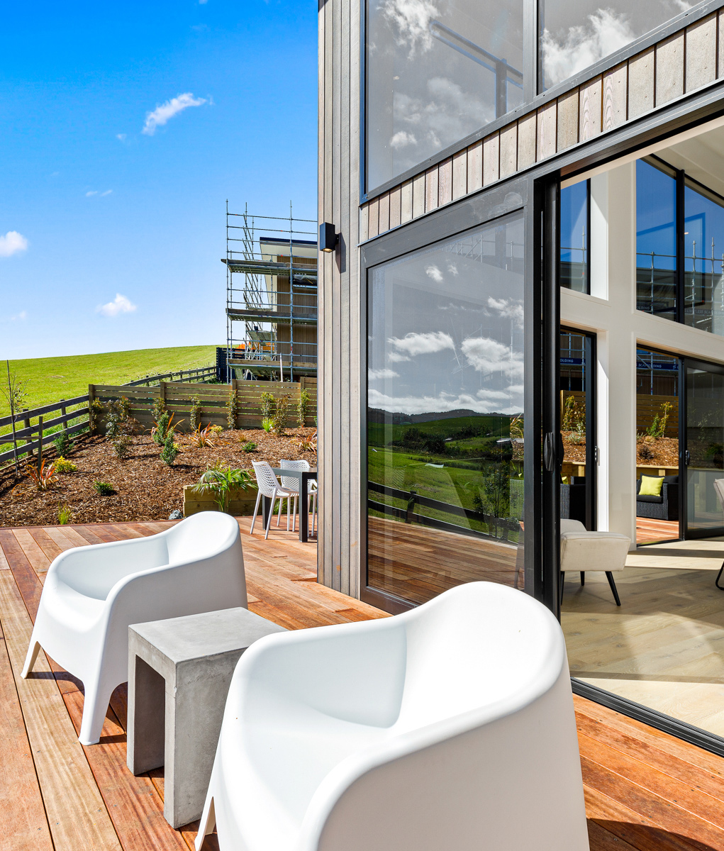 Modern home with large aluminium sliding doors opening to a timber deck with white outdoor chairs and countryside views.