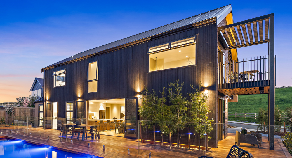 Two-storey home with dark timber cladding, large aluminium windows, and glass balustrades surrounding an illuminated pool at sunset.