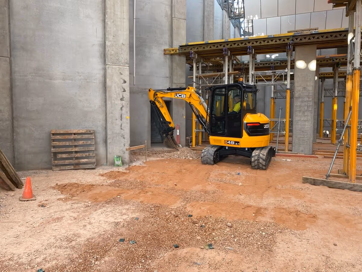A small yellow excavator is parked inside an unfinished building with concrete walls