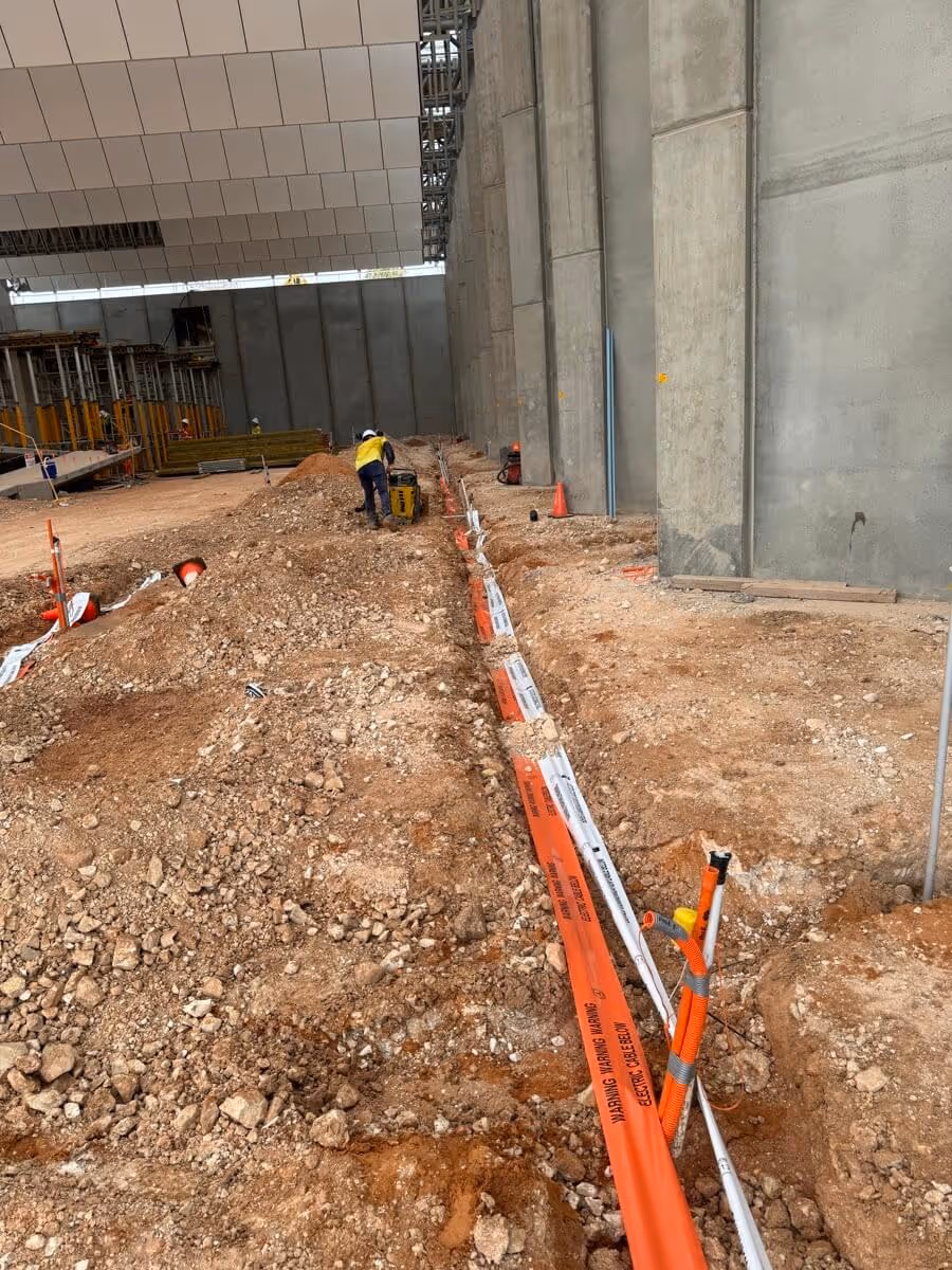 Construction site with a rocky ground and orange barriers. Workers in safety gear are in the background near a large concrete wall, suggesting an indoor project.
