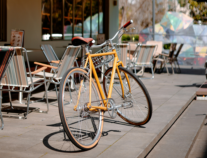 Yellow vintage-style bicycle with a basket parked on a sunlit patio near striped outdoor chairs.