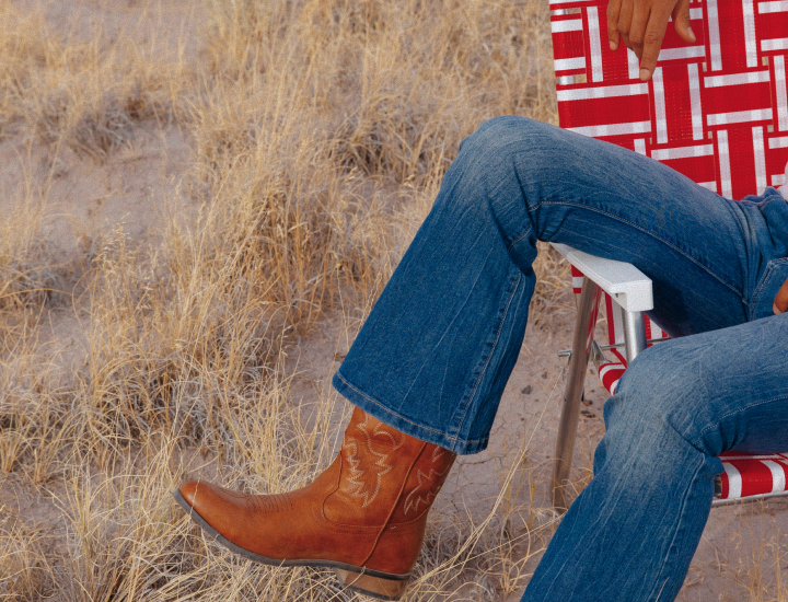 Person in denim jeans and leather cowboy boots sitting in a red-and-white lawn chair in a field.