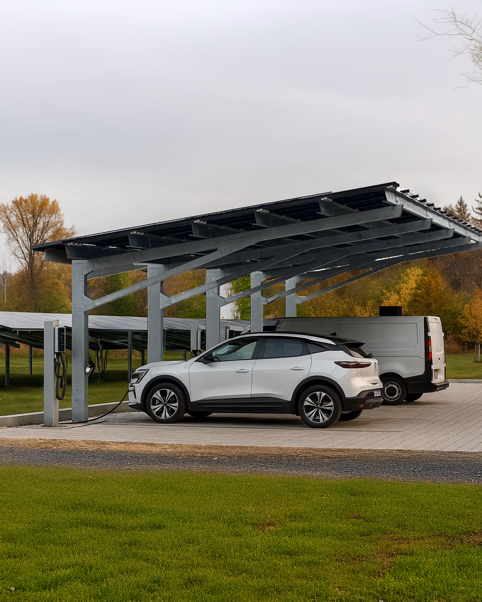 Ein weisser Elektro-SUV lädt unter einem geneigten Solar-Carport aus Metall in halbländlicher Umgebung, neben einem weissen Lieferwagen. Die Konstruktion besteht aus einem verzinkten Stahlrahmen mit Photovoltaikmodulen auf dem Dach. Der Parkplatz ist mit Betonplatten gepflastert und von grünem Gras sowie herbstlich gefärbten Bäumen umgeben – alles unter einem bewölkten Himmel.