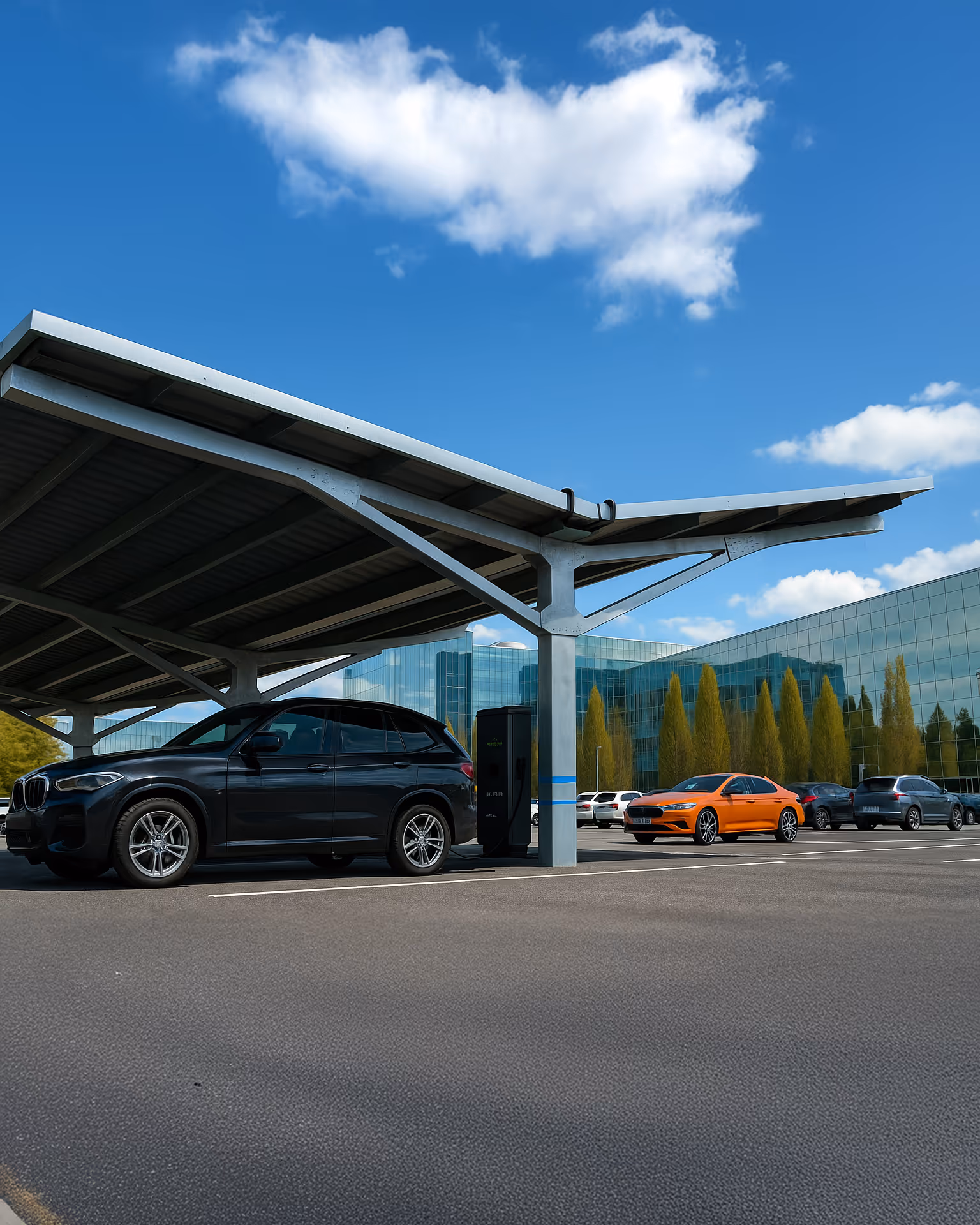 Ein schwarzer SUV lädt unter einem grossen Solar-Carport mit Y-förmiger Stahlkonstruktion auf einem modernen Büroparkplatz. Weitere Fahrzeuge, darunter eine leuchtend orangefarbene Limousine, sind in der Nähe geparkt. Im Hintergrund spiegelt ein verglastes Bürogebäude den blauen Himmel und hohe Bäume wider. Das Wetter ist sonnig mit ein paar verstreuten Wolken.