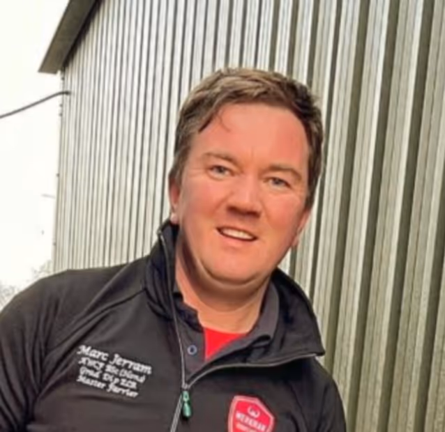 Marc Jerram wearing a black jacket with embroidered text and a red logo standing in front of a corrugated metal wall.