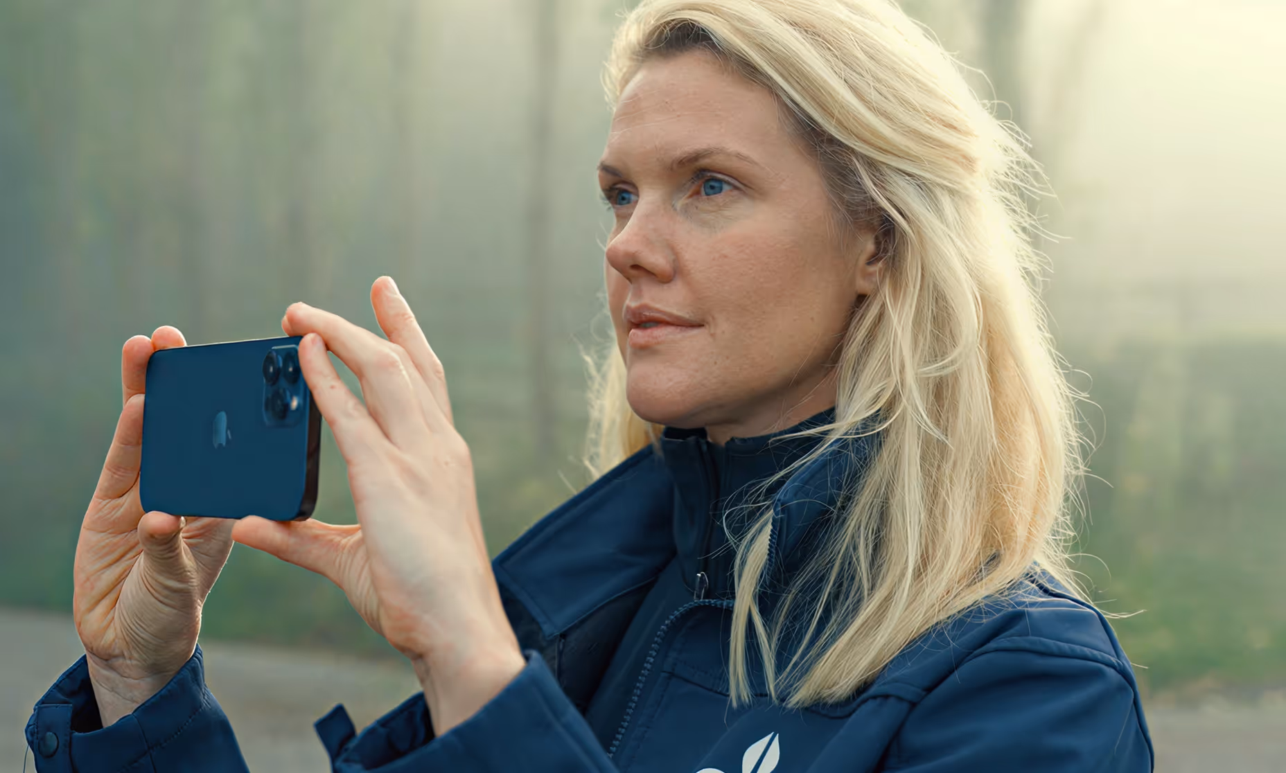 Elin Hernlund in a blue jacket holding a smartphone horizontally, looking intently, outdoors with blurred background.