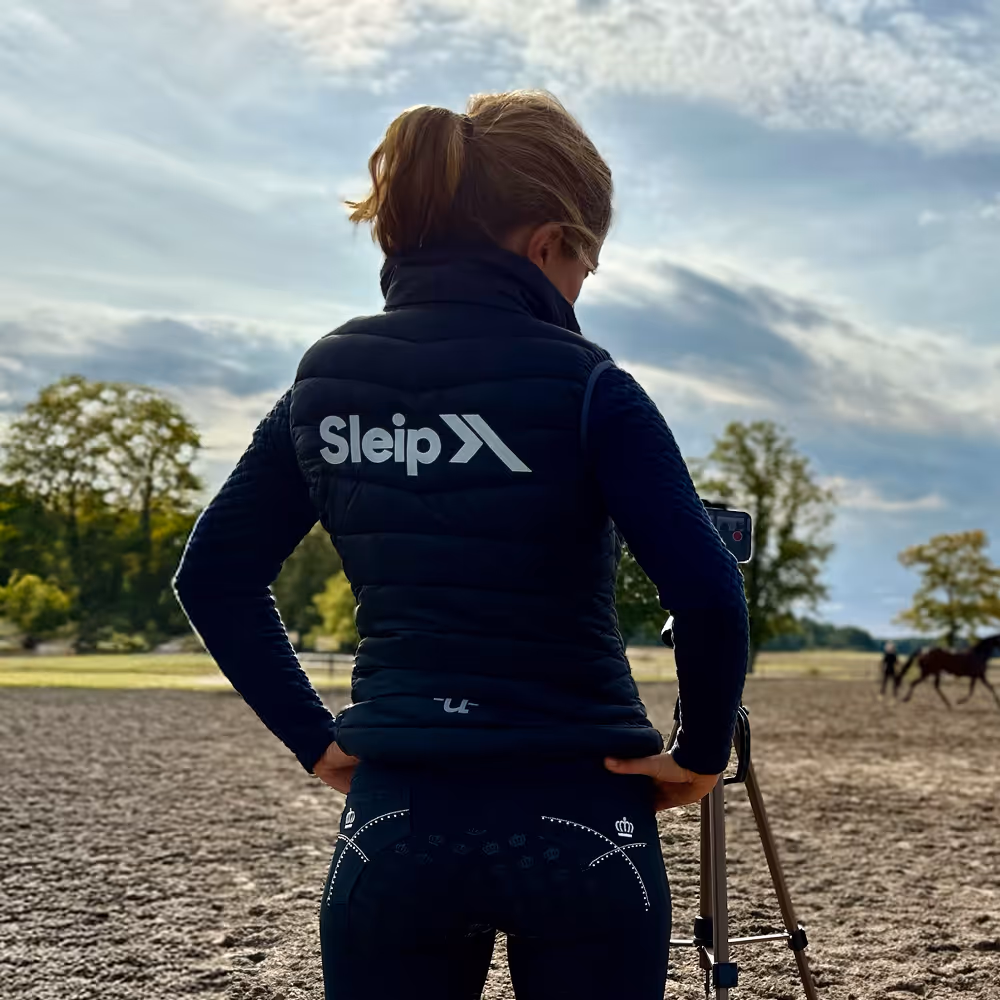 Person wearing a black vest with Sleip logo standing outdoors with hands on hips and a tripod, with horses and trees in the background.