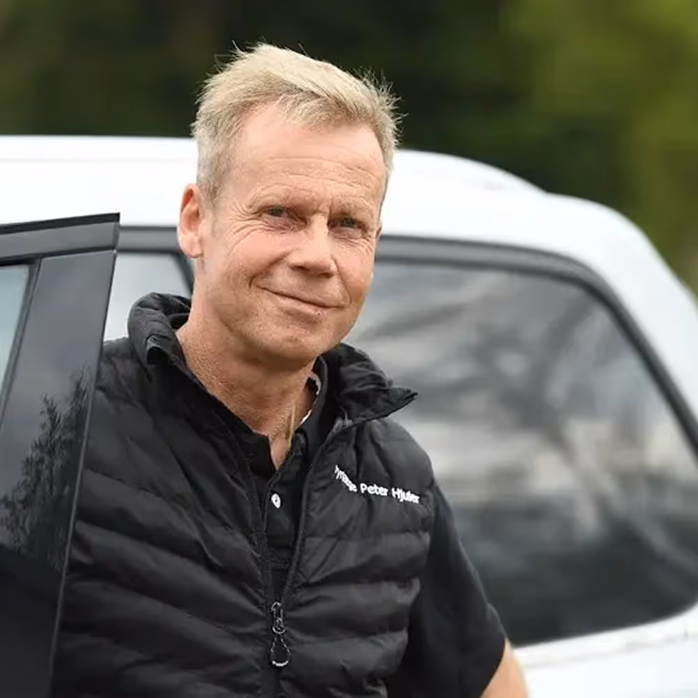 Peter Hjuler wearing black jacket standing next to an open car door with blurred green background.