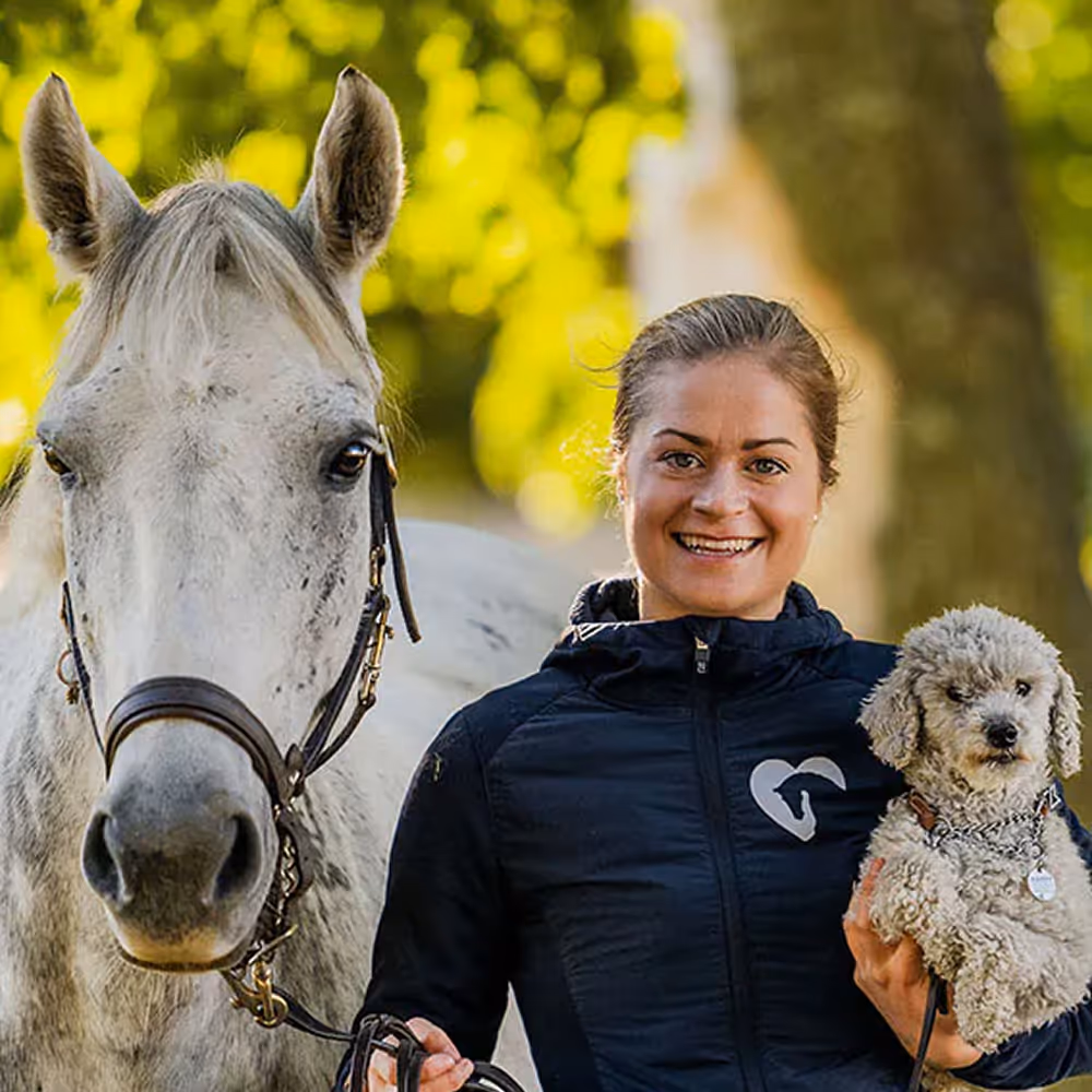 Sara Regårdh holding a small curly-haired dog in one arm and the reins of a white horse with bridle in the other.