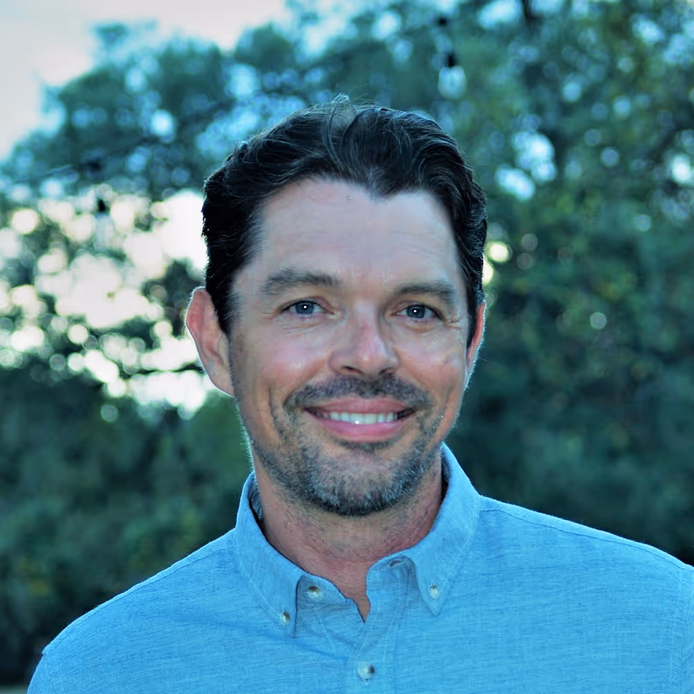 Beua Whitaker with short dark hair and beard wearing a light blue button-up shirt, outdoors with trees in background.